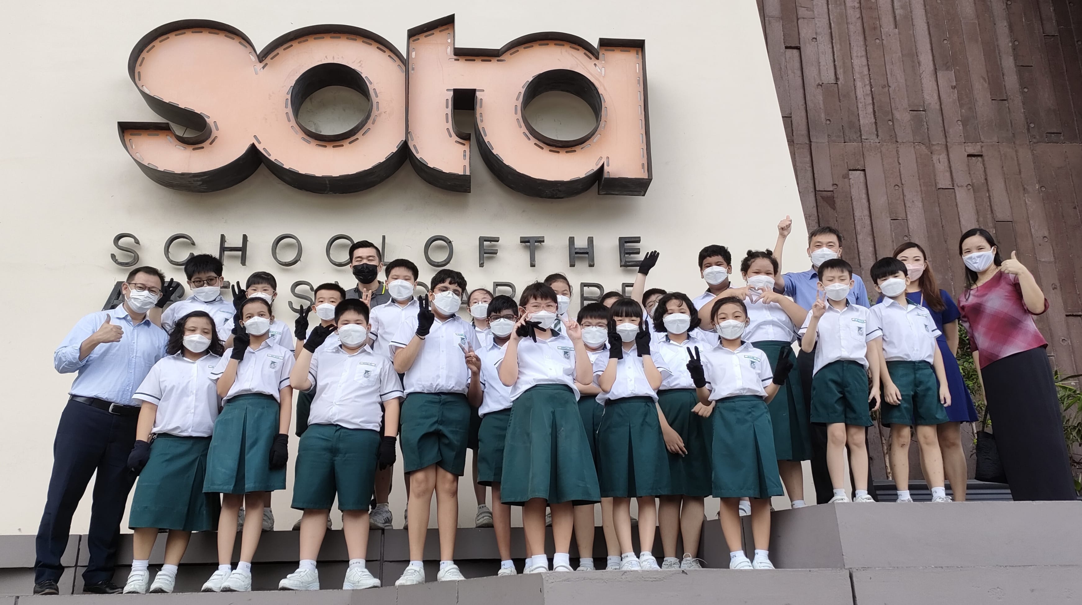 Group of masked students in white and green uniforms posing in front of the SOTA School of the Arts Singapore sign.
